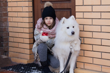A girl in warm clothes hugs a white Samoyed husky dog, a toy red heart in the girl's hand