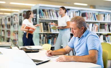 Fototapeta premium Concentrated elderly man writing in planner while searching info in books in the library