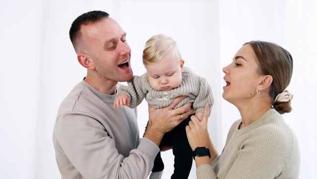 Young family of three in grey sweaters. Caucasian parents hold and kiss their infant son on the cheeks. White backdrop.