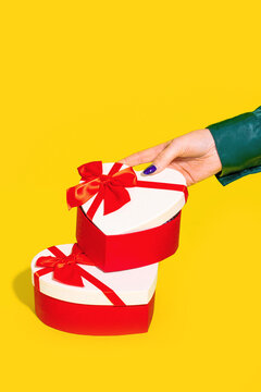 Anonymous hand poised above a stack of heart shaped gift boxes with red ribbons on a yellow background