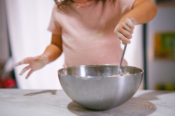 Niña pequeña con harina en las manos removiendo alimento en un bol cromado.