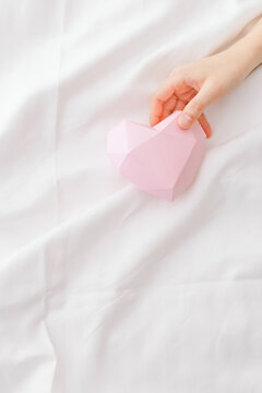 Pink paper valentine heart in woman hand in bed on white crumpled sheet. Minimal style flat lay photo, Valentine's Day, romantic relationship, love, wedding concept, top view, copyspace, pastel