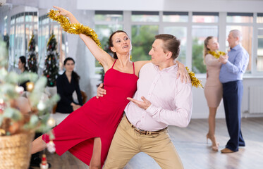 Positive man enjoying romantic waltz with female colleague in red dress and golden tinsel at...