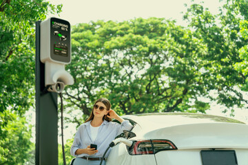 Young woman using smartphone online banking application to pay for electric car battery charging...