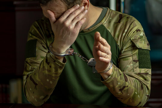 Portrait of a military man in camouflage, hands cuffed, hiding his face.