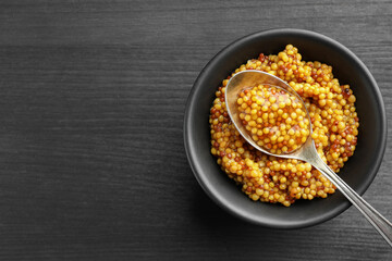 Whole grain mustard in bowl and spoon on black wooden table, top view. Space for text