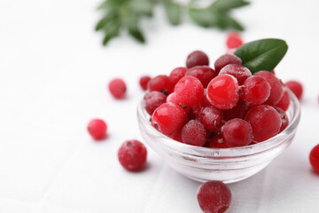 Frozen red cranberries in bowl and green leaves on white table, closeup. Space for text