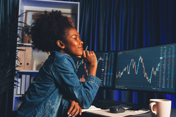Young African woman trader smiling on happy face, looking on screen with valued stock market chart...