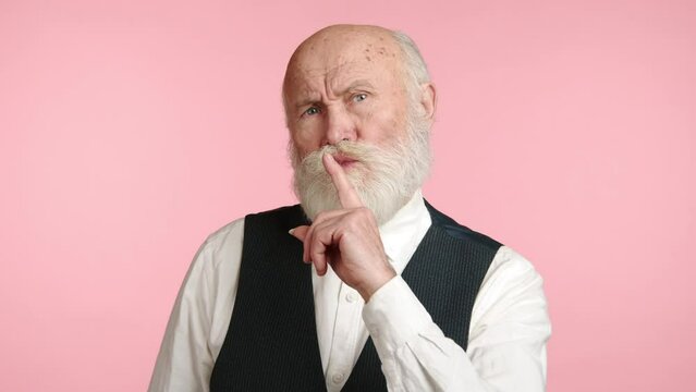 A composed senior stands with his finger pressed to his lips, signaling silence in a playful yet earnest manner, against a backdrop of subtle pink. Camera 8K RAW. 