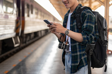 A happy Asian man tourist using his smartphone while standing at a platform in a railway station.