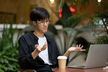 A positive Asian man working remotely at a coffee shop, having an online meeting with his team.