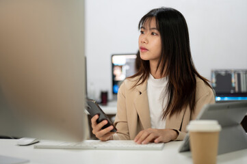 Young beautiful Asian businesswoman working in her agency office, using smartphone and computer.