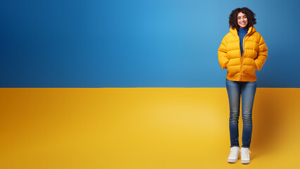 Smiling happy young brunette woman wears yellow puffer jacket, blue jeans and blue turtleneck, both hands in pockets, isolated on split yellow/blue backdrop (Ukrainian flag), copy space, 16:9