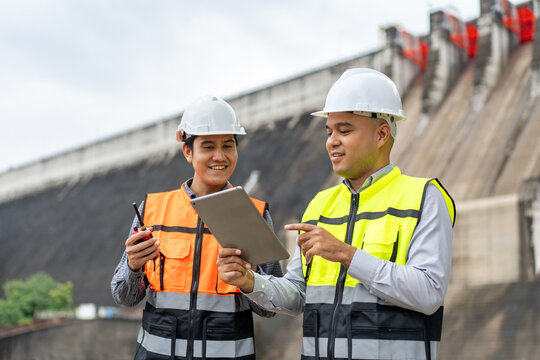 Confident asian two maintenance engineers man inspection discussstion with tablet at construction site dam with hydroelectric power plant and irrigation. Team engineer man working at project