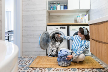 Modern housewife putting dirty linen textile and clothes to washing machine at domestic laundry