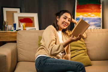 Obraz premium Asian beautiful woman sitting reading book on cozey couch sofa in living room with warm light at night. People female stay home part time relax at home.