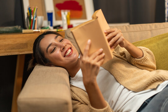 Asian beautiful woman lay down on cozey couch sofa reading book in living room with warm light at night. People female stay home part time relax at home.
