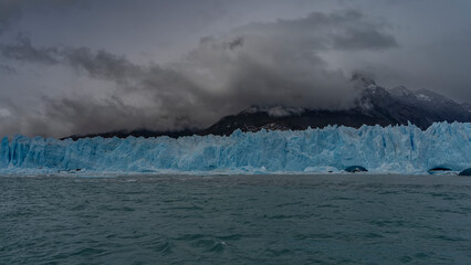 A wall of blue ice with cracks and sharp peaks stretches over a turquoise glacial lake. Melted ice floes float in the water. Mountains in clouds and fog. Perito Moreno glacier. El Calafate. 