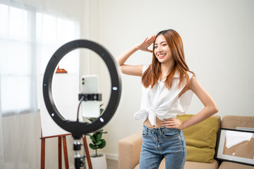 Young trendy influencer asian woman dancing on mobile phone at home in living room. Creator vlogger talent dancing enjoy hobby content recording show video sharing on social media.