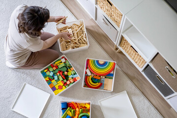 Young woman cleaning childish toy sorting wooden Montessori and plastic details box basket top view