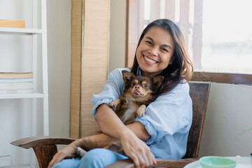 Joyful fit woman sitting having fun with cute dog on the armchair at living room one autumn day , friendship and love for pets.