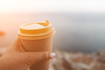 Hand holding Yellow cup with lid, coffee against a backdrop of a blue sky and sea. Illustrating cup...
