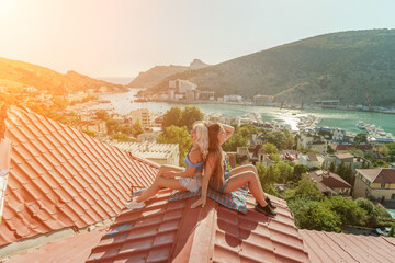 Two women sitting on a red roof, enjoying the view of the town and the sea. Rooftop vantage point. In the background, there are several boats visible on the water, adding to the picturesque scene. - obrazy, fototapety, plakaty