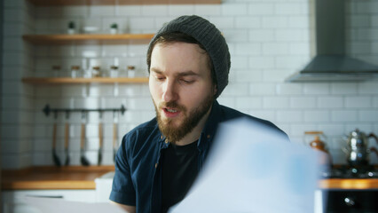 Young angry manager man with beanie sitting in modern kitchen at home, talking, showing reports in...