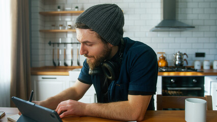 Handsome man with beanie working at home in the kitchen working with his tablet computer, taking...