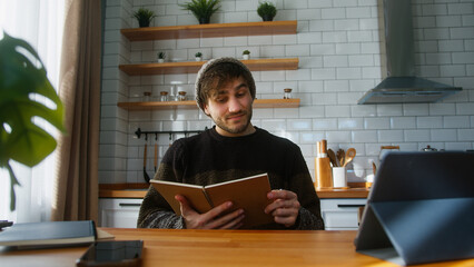Young man student with beanie sitting in modern kitchen at home looking at the notes in notebook he...