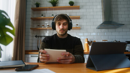 Young man student with beanie sitting in modern kitchen at home wearing headphone listening to...