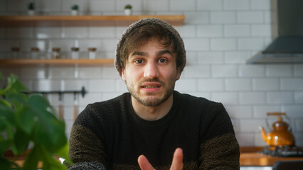 Young adult man with beanie sitting in modern kitchen at home looking to camera having a conference...