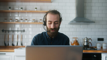 Bearded with hair bun businessman sitting in kitchen wear wireless headset having video conference...