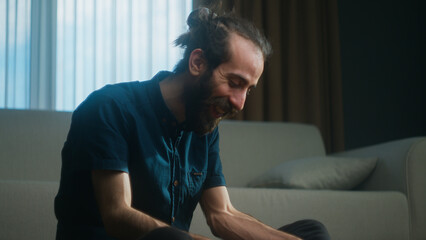 Happy young man with beard and hair in a bun sitting on the floor at home using an app on a tablet...