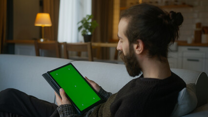 Bearded hair bun man scrolling and tapping center on tablet with green screen mock up display. Male...
