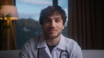 Smiling young professional male doctor wear white uniform with stethoscope having conference video call, chat online with patient, looking camera into webcam	
