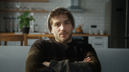 Bored young man with beanie sitting on sofa at home with kitchen background, having a conference...