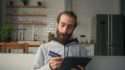 Young man with hair bun sitting on sofa at home with kitchen background, entering credit card...