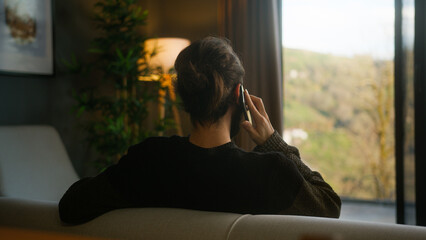 Back view of bearded man with his hair in a bun talking on his mobile phone, sitting on the sofa at...