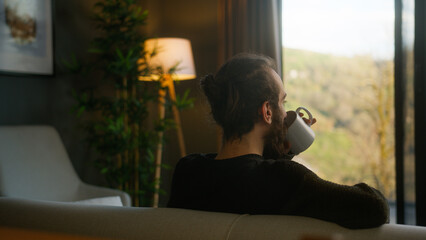 Back view of bearded man with his hair in a bun sitting on the sofa at home, relaxing and drinking...