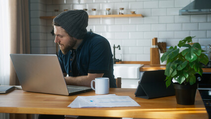 Focused man with beanie working at home in the kitchen working with his laptop taking notes, doing research. Busy freelancer working on modern tech notebook
