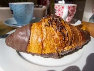 Detail shot of a breakfast with a large chocolate croissant and two coloured coffee cups on a white table.