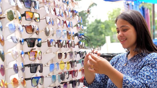 A pretty Indian girl trying out colorful sunglasses in the market - street side shopping  local bazaar  local market. A young girl doing shopping - fashion conscious college going girl  trendy fash...