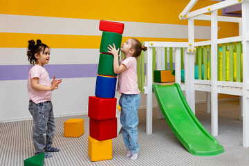 Two girls play with colorful, large, soft blocks-constructor for kindergarten. A fun game and developing skills in building castles and houses. Happy children have fun in the children's room.