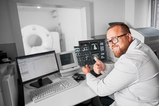 Medical computed tomography or MRI scanner. Doctor sitting at computer. Male specialist wearing glasses and white robe, looking at camera, smiling, showing thumbs up. Concept of modern diagnostics.