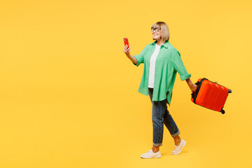 Traveler elderly woman wear green casual clothes hold mobile cell phone bag isolated on plain yellow background. Tourist travel abroad in free spare time rest getaway. Air flight trip journey concept. - obrazy, fototapety, plakaty