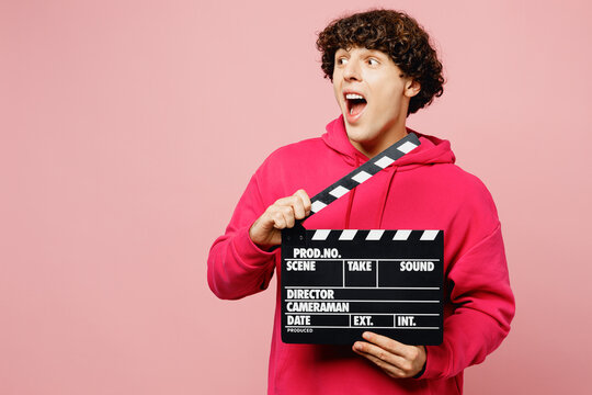 Young surprised excited Caucasian man wear hoody casual clothes : hold in hand classic black film making clapperboard isolated on plain pastel light pink background studio portrait. Lifestyle concept - Powered by Adobe