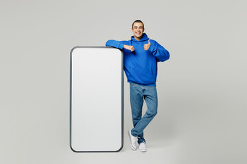 Full body happy young middle eastern man wear blue hoody casual clothes big huge blank screen mobile cell phone with workspace copy space area show thumb up isolated on plain solid white background