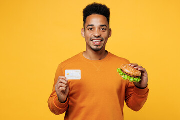 Young happy man wear orange sweatshirt casual clothes eat burger hold credit bank card isolated on...