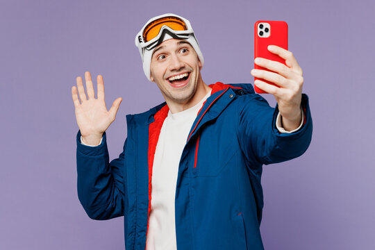 Skier man wear warm blue windbreaker jacket ski goggles mask hat do selfie shot on mobile cell phone waving hand spend extreme weekend winter season in mountains isolated on plain purple background.